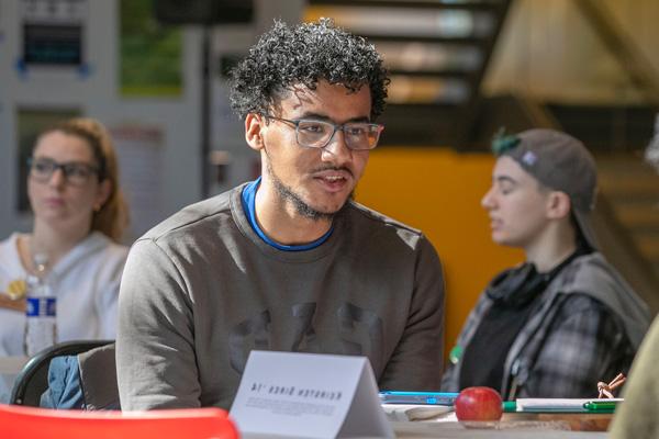 male college student sits at a table in discussion with another student